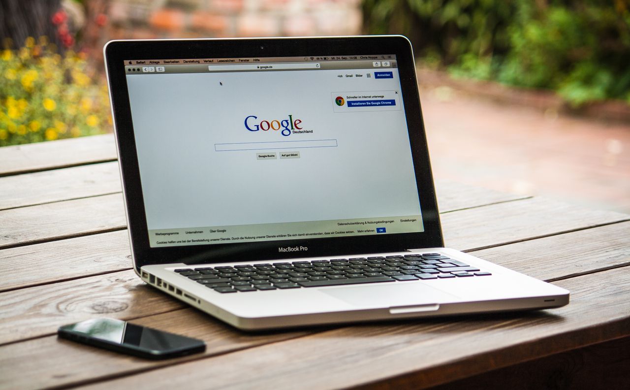 A MacBook Pro showing Google Search on a wooden table outside, beside a smartphone.